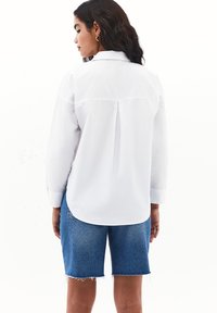 Camisa blanca de manga larga con cuello abotonado, detalle de pliegue en la espalda y dobladillo redondeado, combinada con pantalones cortos de denim azul.