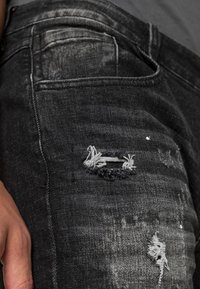 Czarne dżinsy z denimu z przetarciami, posiadające postrzępioną dziurę i subtelne wyblaknięcia. Kieszeń widoczna po prawej stronie.