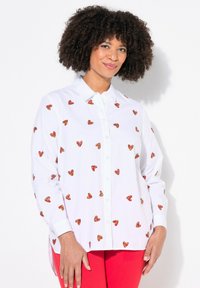 Camicia bianca con ricami a forma di cuore rossi e arancioni sparsi, maniche lunghe e orlo leggermente curvato. Indossata con pantaloni rossi.