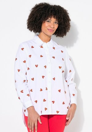 Camicia bianca con ricami a forma di cuore rossi e arancioni sparsi, maniche lunghe e orlo leggermente curvato. Indossata con pantaloni rossi.