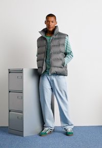 Grå puffervest over en grønstribet langærmet skjorte, med lyseblå denim, stående ved siden af et gråt arkivskab og iført grønne sneakers.