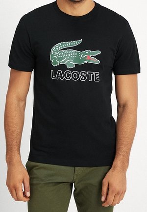 Mand iført sort T-shirt med en stor grøn krokodillelogo og ordet "LACOSTE" trykt nedenunder, kombineret med olivengrønne bukser.