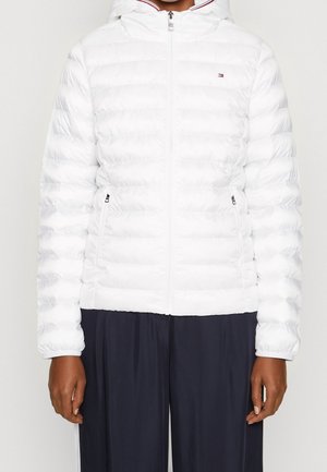 Veste blanche matelassée à capuche avec fermeture éclair à l'avant et poches latérales, portée avec un pantalon large foncé, montrant le haut du corps et les bras.