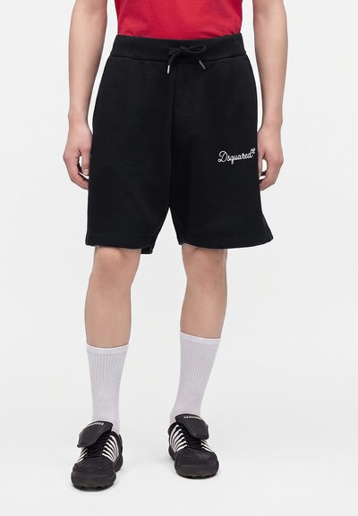 Personne portant un short noir à cordon avec le logo blanc "Dsquared²", des chaussettes blanches montantes et des chaussures de sport noires avec languettes cachées.