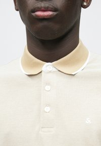 Gros plan d'une personne portant un polo beige avec un col marron clair et trois boutons blancs, montrant une partie du cou et des lèvres.
