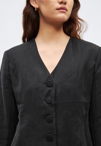 Čierne ľanové sako s výstrihom do V, piatimi guľatými gombíkmi a textúrovanou látkou. Akcenty zahŕňajú priliehavý strih a hladké rukávy.