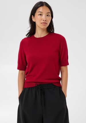 Jeune femme aux cheveux noirs portant un haut rouge à manches courtes et un pantalon noir avec cordon, debout devant un fond clair uni.