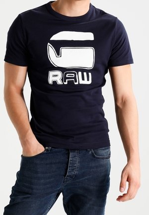 Muž v námořnickém modrém tričku s velkým bílým logem velryby a nápisem "RAW" vytištěným níže, doplněný o tmavě modré džíny.