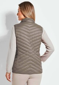 Padded Weste in Taubengrau mit Zickzack-Steppung, hohem Kragen und leichtem Design. Darunter ist ein weiches beiges Langarmshirt zu sehen.