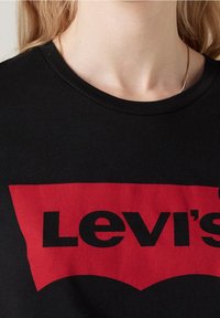 Camiseta de algodón negra con un prominente logo rojo de Levi's con una forma curva distintiva y letras negras en negrita en la parte delantera.