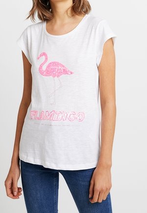 Kobieta w białej koszulce z krótkim rękawem z różowym flamingiem z cekinów i napisem "FLAMINGO", w połączeniu z niebieskimi dżinsami.