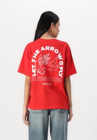 Rød oversized t-skjorte med et stort hvitt grafisk trykk av en flyvende cherub og teksten "LET THE ARROWS FLY LVRS CLUB," laget av bomull.