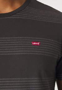 T-shirt noir à col rond avec de fines lignes de couture horizontales blanches et un petit logo Levi's rouge sur la poitrine.