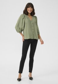 Blusa verde oliva con maniche a sbuffo, scollo a V e dettagli con bottoni. Presenta un bordo in pizzo lungo i bordi. Abbinata a pantaloni neri aderenti e scarpe nere.