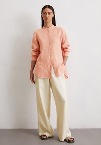 Perzikkleurig linnen shirt met een mandarin kraag, knoopsluiting aan de voorkant en lange mouwen, gecombineerd met brede witte linnen broek en donkere sandalen.