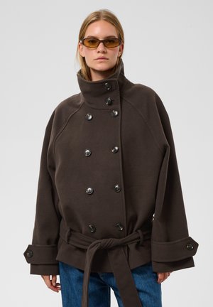 Femme portant un manteau oversize marron foncé à double boutonnage avec un col haut et une ceinture nouée, un jean bleu et des lunettes de soleil teintées ambre.
