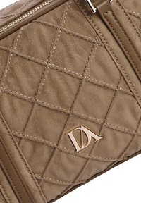 Bolso acolchado beige con costuras diagonales, detalles de cuero suave y un logo dorado. Cierre de cremallera en el borde superior.