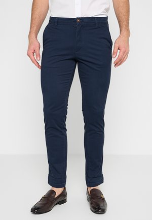 Slim-fit marineblauwe broek gedragen met bruine leren loafers en een witte shirt, getoond van taille tot voeten.