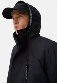 Marineblå vandtæt jakke med høj krave, justerbar hætte og lynlåslommer. Har en glat tekstur og minimalistisk design.