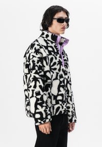 Zwart-witte pluche fleece jacket met abstracte patronen, voorzien van een lavendelknopenkraag. Model draagt zwarte zonnebril.