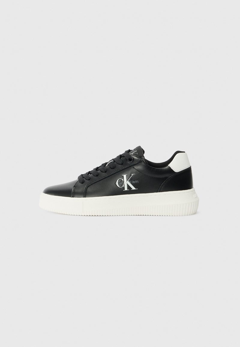 Sneaker basse in pelle nera con suola in gomma bianca, punta rotonda, lacci piatti e dettaglio del logo impresso sul lato. Accento sul tallone in contrasto.