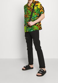 Kurzarmhemd in hellgrün und orange mit tropischen Mustern, kombiniert mit schwarzen, verwaschenen Jeans und schwarzen Rutschen-Sandalen.
