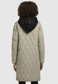 Abrigo largo acolchado de tela beige con capucha negra que presenta texto en relieve. El diseño incluye un corte holgado y bolsillos laterales.