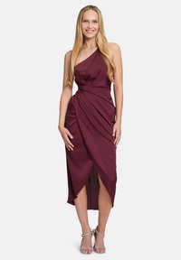 Robe bordeaux à une épaule avec un tissu drapé, longueur mi-mollet, dotée d'une fente sur le côté et d'une texture lisse. Associée à des sandales à talons argentées.
