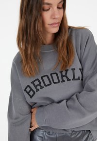 Gray sweatshirt with "BROOKLYN" in black embroidery; features a crew neckline and relaxed sleeves, made of a soft fabric.
