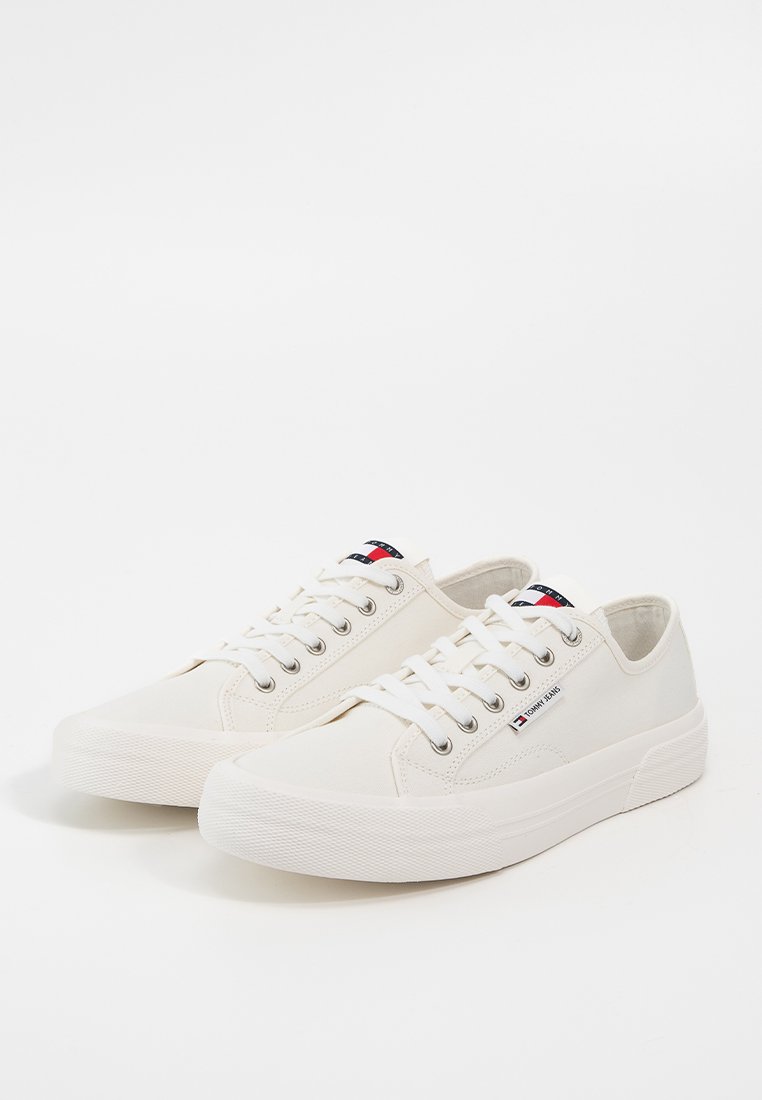 Zapatillas de lona blancas con suela de goma, punta redonda, diseño con cordones y pequeño detalle de logo en el lateral. Textura suave y estilo minimalista.