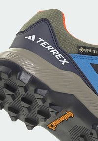 Niebiesko-szary but outdoorowy z fakturowaną cholewką, pomarańczowymi akcentami oraz wytrzymałą czarną podeszwą z wyraźnym bieżnikiem. Widoczne logo marki.