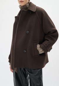 Abrigo de lana marrón, de doble botonadura con grandes botones negros, bolsillos laterales y un corte relajado. Cuello de tortuga de punto visible debajo. Pantalones negros.