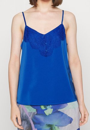 Haut bleu sans manches avec détail en dentelle et boutons porté par une personne, associé à une jupe avec un grand motif floral bleu et vert.