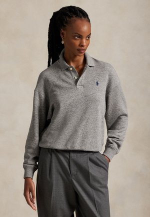 Une femme porte un polo gris ample à manches longues, orné d’un petit logo sur la poitrine, avec un pantalon gris foncé, sur un fond beige uni.