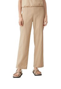 Beige Schlaghosen, kombiniert mit beigen Sandalen mit überkreuzten Riemen und flachen schwarzen Sohlen, gezeigt von der Taille bis zu den Füßen.