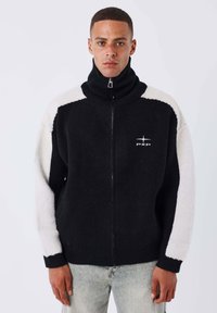 Homme portant un sweat à zip noir avec des manches blanches et un col haut, se tenant devant un fond blanc uni.