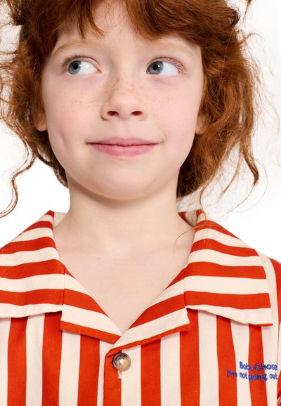 Jeune enfant roux avec des taches de rousseur portant un t-shirt rayé rouge et blanc, regardant sur le côté avec un léger sourire.