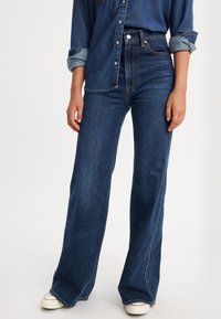 Femme portant un jean large taille haute bleu foncé, une chemise en denim aux manches retroussées et des baskets blanches sur un fond uni.
