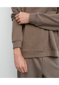 Ruda medvilninė palaidinė su gumuotomis rankovėmis ir apačia, pasižyminti laisvu siluetu ir tekstūruota medžiaga. Rankos laikoma rankovė, demonstruojanti detalę.