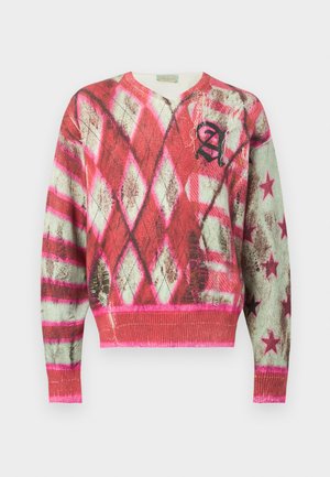 Mann som har på seg en rosa og hvit mønstret genser med svarte bokstaver, kombinert med falmete grafiske jeans, svart belte og svarte joggesko.