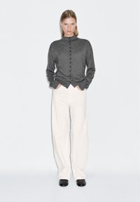 Gilet gris à boutons avec un col haut, des manches longues et des boutons noirs, associé à un pantalon large blanc et des chaussures noires.