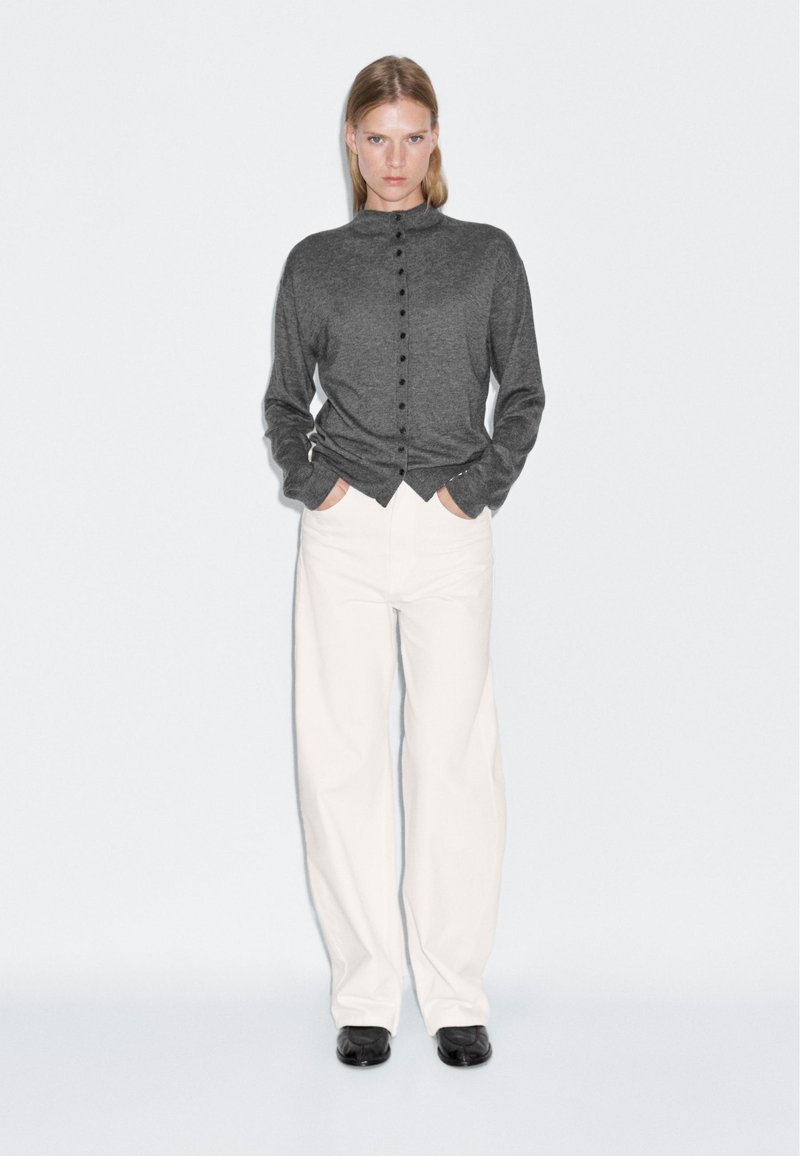 Gilet gris à boutons avec un col haut, des manches longues et des boutons noirs, associé à un pantalon large blanc et des chaussures noires.