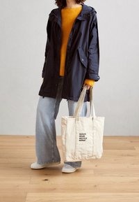 Personne portant une veste marine, un pull jaune, un jean large, des chaussures blanches, tenant un sac fourre-tout beige avec le texte "RÉDUIRE RÉUTILISER RECYCLER RÉPÉTER".