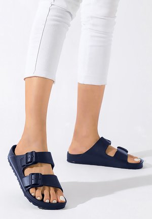 Voeten met marineblauwe sandalen met dubbele gespen, gecombineerd met cropped witte broeken op een witte achtergrond.