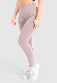 Leggings a vita alta di un leggero colore malva, realizzati in un tessuto liscio e elasticizzato. Caratterizzati da una cintura larga e un design senza cuciture. Indossati con scarpe da ginnastica bianche.