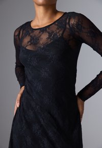 Robe noire avec de longues manches en dentelle transparente, motif floral, corsage ajusté et superposition texturée, créant un effet visuel en couche.