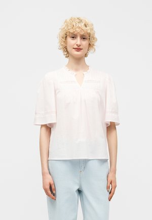 Blondynka w luźnej białej bluzce z bufkowymi rękawami i jasnoniebieskich dżinsach, stojąca na jednolitym tle.