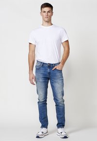 Jeans de denim azul con un corte Slim, ligero desgaste y un diseño clásico de cinco bolsillos, combinados con una camiseta blanca de algodón lisa y zapatillas.