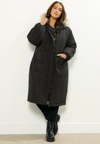 Musta pitkä takki, joka on valmistettu sileästä kankaasta ja jossa on huppu, jossa on tekoturkistelaus, elastinen vyötärö ja kaksi suurta etutaskua.