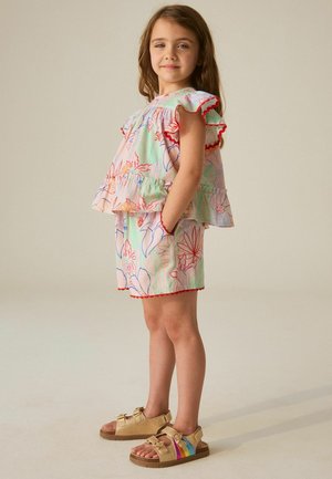 Mladé dievča stojí s rukami v kapse, oblečená v kvetovaný top a šortky s červeným lemom a béžové sandále s dúhovými remienkami.